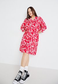 Κόκκινο φόρεμα με λευκό αφηρημένο σχέδιο καρδιάς, τρεις τέταρτα μ�ανίκια, ζώνη στη μέση, σε συνδυασμό με μαύρα ψηλά sneakers και λευκές κάλτσες.