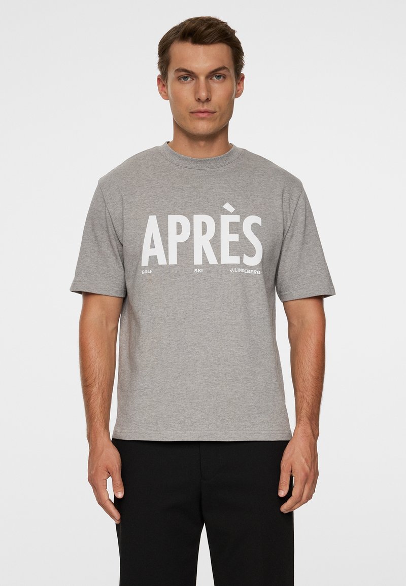 Mężczyzna w szarej koszulce z krótkim rękawem z białym napisem "APRÈS" oraz mniejszymi słowami "Golf", "Ski" i "J. Lindeberg" z przodu.