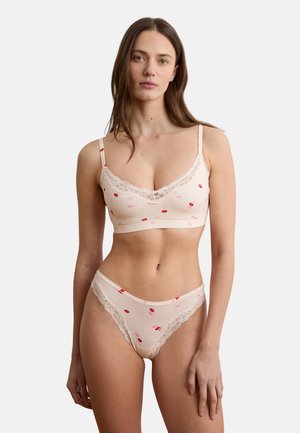 Vrouw draagt een lichtroze lingerieset met rood-roze lippenafdrukken en kanten randen, staand tegen een effen witte achtergrond.