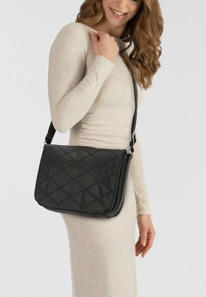 Gepolsterte schwarze Leder-Umhängetasche mit schlankem Profil, verstellbarem Gurt und Akku mit silbernen Hardware-Details, getragen über einem cremefarbenen Kleid.