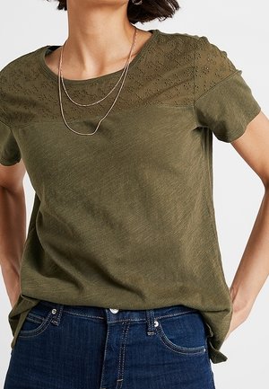 Olivgrünes T-Shirt mit kurzen Ärmeln und floraler Lochstickerei am Passe, weicher Stoff, entspannte Passform, kombiniert mit dunkelblauen Jeans.