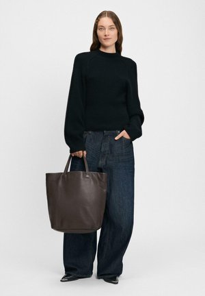 Vrouw in een zwarte trui en wijde donkere jeans, met een grote donkerbruine leren draagtas met twee handvatten.