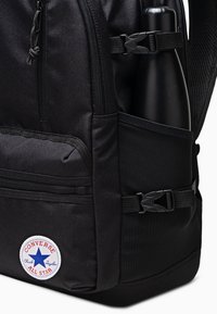 Mochila de tela negra con múltiples compartimentos con cremallera, bolsillo de malla lateral para una botella de agua y un parche circular del logo de Converse.