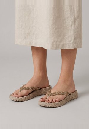 Feet wearing beige woven platform flip-flops with beaded straps, standing on a light floor with a beige linen skirt.
