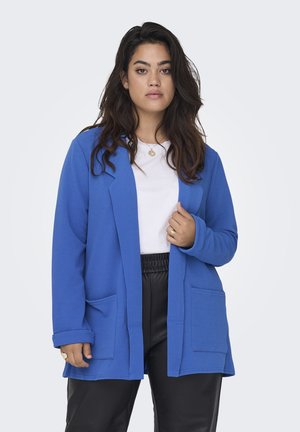 Cardigan largo azul con acabado texturizado, de frente abierto y dos bolsillos frontales, combinado con una camisa blanca y pantalones negros.