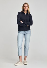 Svart zip-up sweatshirt med ribbade manschetter, ljusblå cropped jeans och beige sneakers. Modellen står med armarna i kors mot en neutral bakgrund.