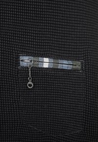 Schwarzer, strukturierter Stoff mit einer Reißverschlusstasche, die mit einem gestreiften Reißverschlussrand in Blau, Weiß und Grau verziert ist, sowie einem kleinen Kettenanhänger mit einem hexagonalen charm.