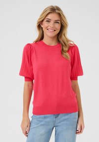 Pull en tricot corail avec des manches courtes à volants côtelés et un col arrondi. Texture douce, coupe ajustée, associé à un jean en denim bleu clair.
