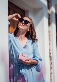 Light blue, flowing dress with a v-neck and gathered waist; holding a small pink wallet; wearing black sunglasses. Soft texture.