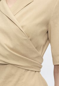 Μπεζ ένδυμα σε στυλ τύλιγμα με μανίκια τριών τετάρτων, εφαρμοστή μέση και εμφανείς ραφές σε λείο ύφασμα.