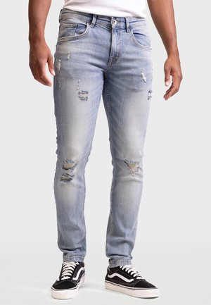 Homme portant un jean slim déchiré bleu clair et des baskets noires et blanches, debout sur un fond blanc.