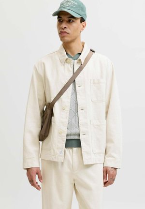 Mężczyzna ubrany w kremową kurtkę i spodnie, wzorzysty sweter, zielona czapka i brązowa torba na ramię, stojący na tle jednolitego jasnego tła.