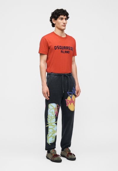 Άνδρας με κόκκινο t-shirt Dsquared2 Milano, μαύρο παντελόνι φόρμας με πολύχρωμα σχέδια graffiti και σκοτεινά αθλητικά παπούτσια, στέκεται μπροστά από απλό φόντο.