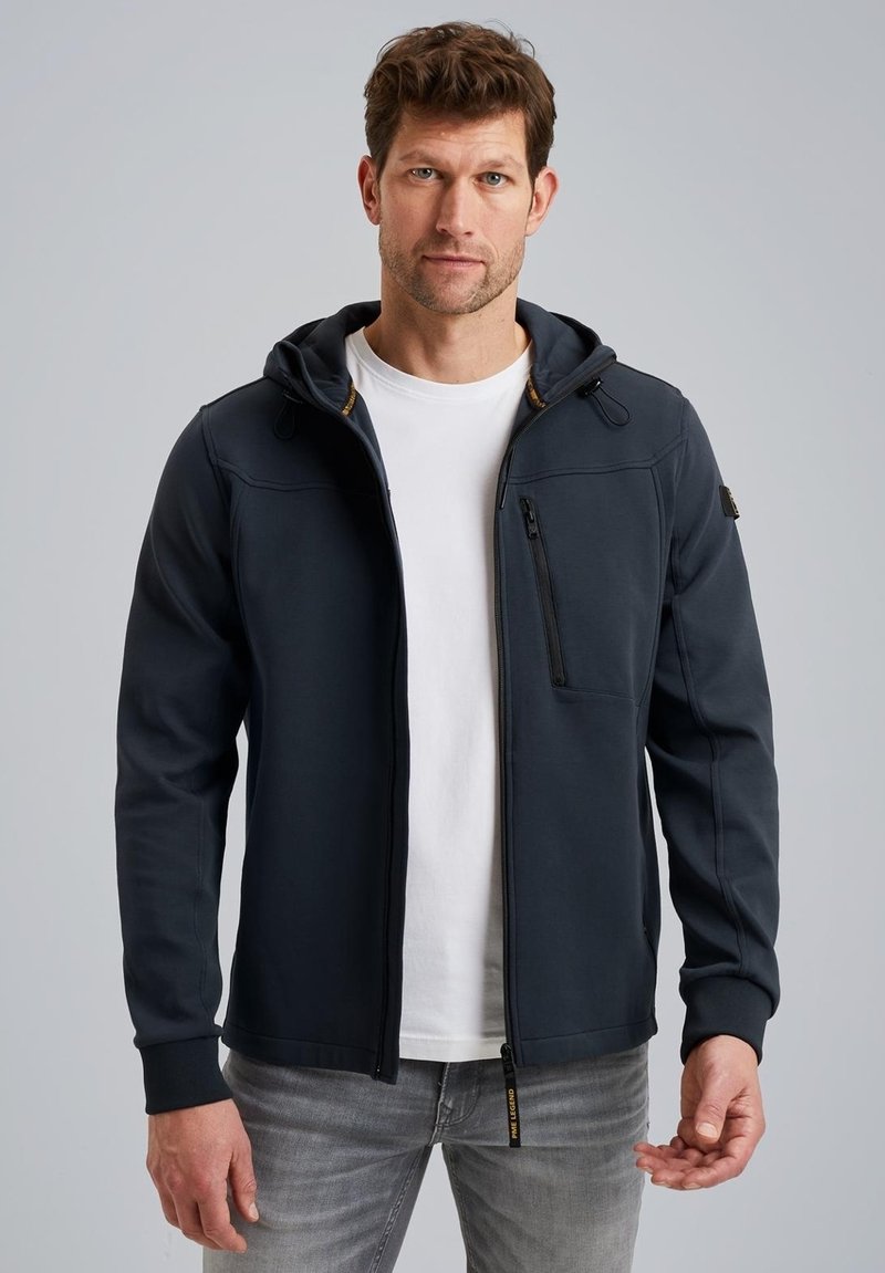 Sweat à capuche zippé bleu marine foncé avec une texture lisse, capuche réglable et poche zippée à l'avant, associé à un t-shirt blanc et un jean gris.