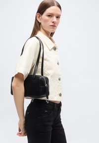 Νεαρή γυναίκα που φοράει κρεμ κοντομάνικο πουκάμισο με κουμπιά και μαύρο τζιν, κρατώντας μια μαύρη δερμάτινη τσάντα ώμου.