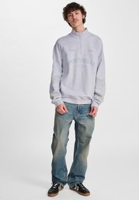 Ljust grå sweatshirt med halv dragkedja, texten "SPORTSWEAR", kontrasterande grå ränder på ärmarna, kombinerat med blekta blå jeans och svarta sneakers.