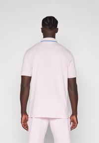 Camisa polo rosa claro com um destaque de colarinho azul, mangas curtas e uma bainha ventilada, feita de um tecido suave e leve. Vista traseira.