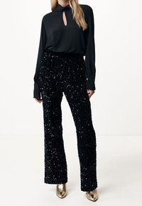 Blusa nera a maniche lunghe con scollatura a chiave abbinata a pantaloni larghi neri con paillettes e scarpe con tacco dorate, su uno sfondo bianco.