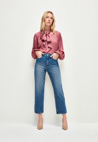 Blusa rosa con lazo y detalles de volantes, combinada con jeans de mezclilla azul de tiro alto. La modelo lleva tacones beige. Tejido suave y corte relajado.