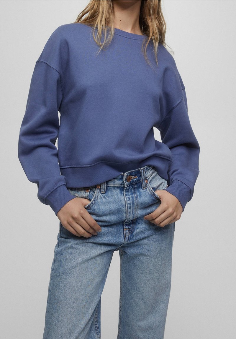 Personne aux longs cheveux portant un sweat-shirt bleu et un jean taille haute bleu clair avec les mains dans les poches, sur un fond uni.