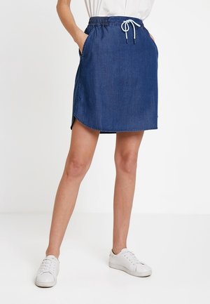 Kvinde iført en blå denim nederdel med elastisk talje og hvide snore, kombineret med hvide sneakers, stående med hænderne i lommerne.