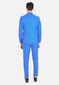 Abito blu con finitura testurizzata, con uno spacco singolo sul retro, tagliato in una vestibilità su misura, abbinato a scarpe nere.