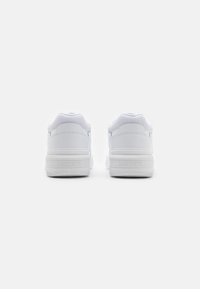 Fehér sportcipők sima bőr felsőrésszel, hálós betétekkel, párnázott nyakkal és mintás gumitalppal. Lacoste felirat a saroknál.