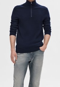 Man iförda en marinblå halvpolo med ribbad mudd och blekta ljusblå jeans, med ena handen i fickan, mot en neutral bakgrund.