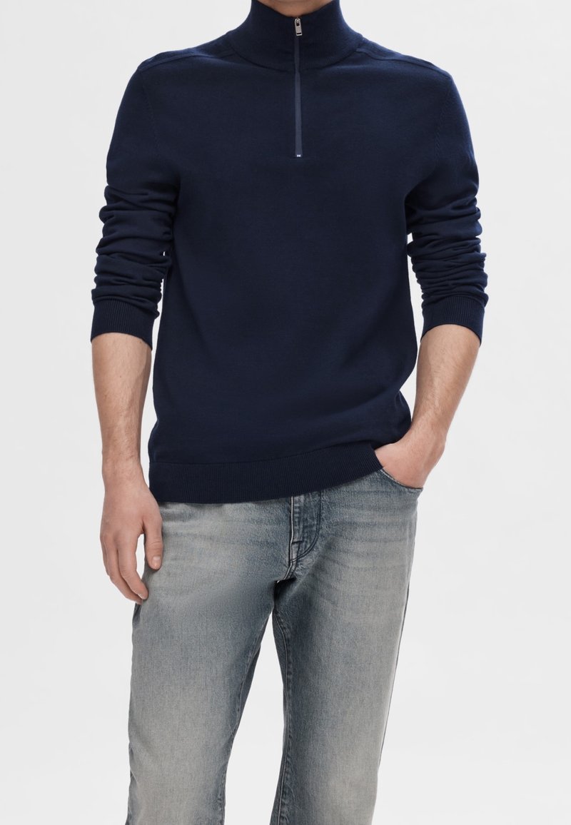 Man iförda en marinblå halvpolo med ribbad mudd och blekta ljusblå jeans, med ena handen i fickan, mot en neutral bakgrund.