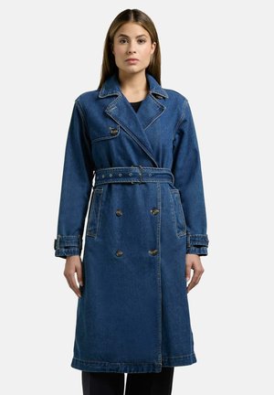 Kvinne som bærer en lang, mørk blå denim trenchcoat med dobbeltradede knapper og belte i midjen, stående mot en ensfarget bakgrunn.