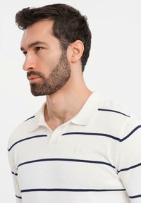 Muž s vousy a tmavými vlasy, oblečený v bílém polo tričku s dlouhým rukávem a námořnickými vodorovnými pruhy, dívající se vlevo na jednobarevném pozadí.