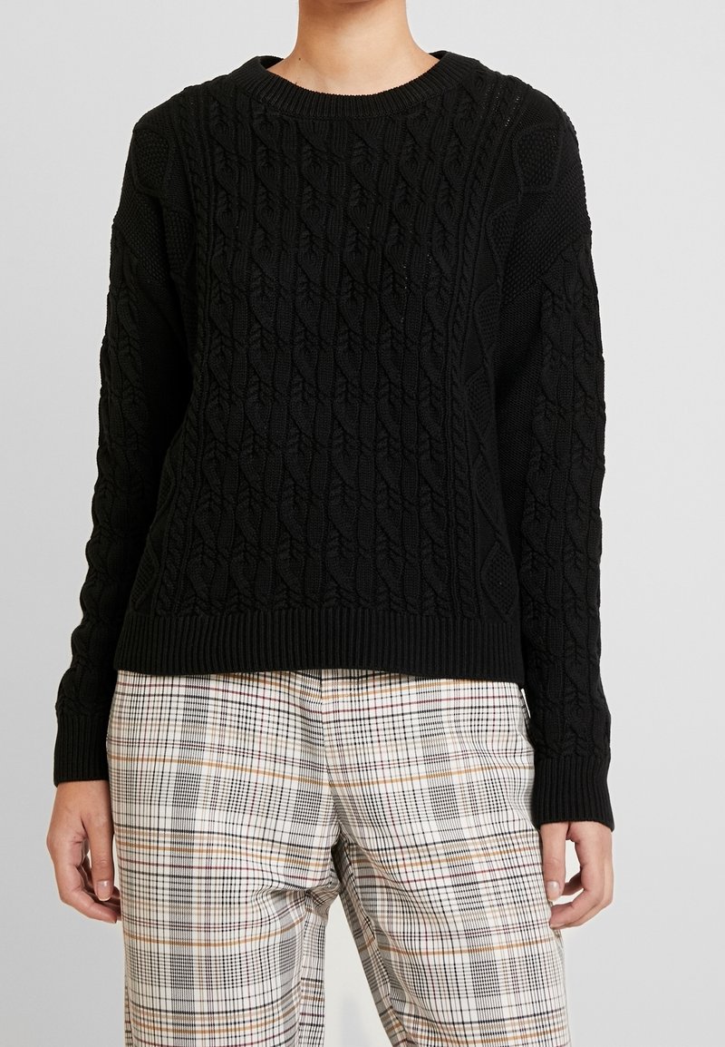 Personne portant un pull noir en maille torsadée avec poignets et bas côtelés, associé à un pantalon à carreaux clair avec des lignes orange et noires.
