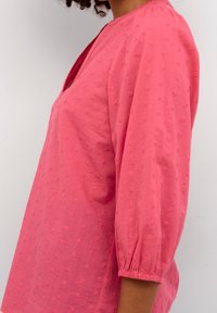 Donna che indossa una blusa rosa con texture e maniche a sbuffo a tre quarti su uno sfondo bianco semplice.
