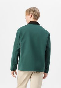 Grøn stofjakke med brun krave. Har en afslappet pasform, lange ærmer og knapmanchetter. Glat tekstur, minimalistisk design.
