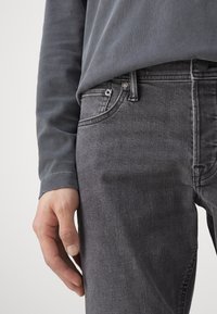 Grå langermet skjorte lagd over mørkegrå denimjeans. Buksene har en klassisk fem-lommers design med synlig søm og maskinvare.