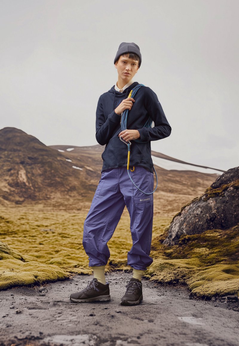 Abbigliamento outdoor: felpa blu navy, pantaloni viola, berretto grigio e scarpe da trekking marroni, con una corda da arrampicata in mano. Sfondo con paesaggio texturizzato.