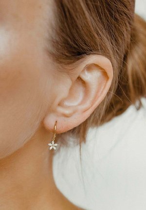 Goldene Creolen mit floralen Designs, die aus fünf Blütenblättern und kleinen Edelsteinen bestehen, hängen vom Ohrläppchen vor einem hellen Hintergrund.