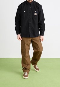 Schwarzes Button-up-Hemd mit zwei Brusttaschen, kombiniert mit braunen Cargo-Hosen mit Seitentaschen und braunen Sneakers mit weißen Sohlen.