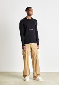 Maglione nero a maglia con scollo rotondo e logo bianco "Calvin Klein Jeans", abbinato a pantaloni cargo beige e sneakers bianche.