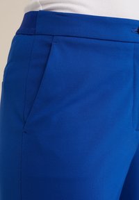 Modré elastické kalhoty s plochým předním dílem, bočními kapsami a hladkým pásem v pase, se zdobeným střihem a moderním designem.