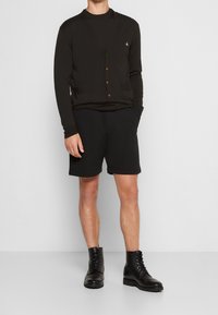 Svart cardigan med lång ärm, knäppning fram och två fickor över svarta shorts. Bärs med mönstrade svarta läderstövlar.