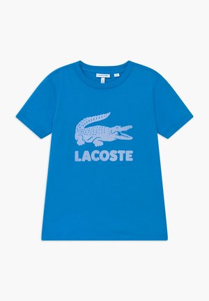 Niebieska koszulka z krótkim rękawem z dużym jasnoniebieskim krokodylem i napisem "LACOSTE" umieszczonym centralnie z przodu.