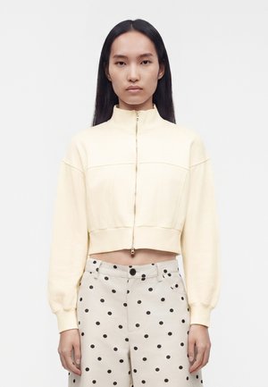 Kvinde med langt mørkt hår iført en cremefarvet cropped jakke med lynlås og højtsiddende beige bukser med sorte polkaprikker, stående foran en ensfarvet baggrund.