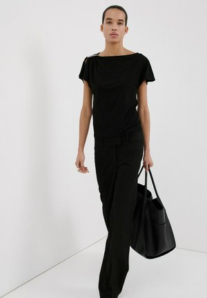 Person trägt komplett schwarzes Outfit mit kurzärmeligem Oberteil und weitem Hosenbein, hält große schwarze Tragetasche und steht vor weißem Hintergrund.