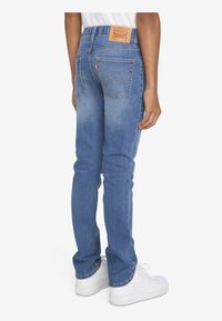 Jeans de mezclilla azul con corte recto, bolsillos traseros y un parche de cuero marrón en la cinturilla. Llevados con zapatillas blancas.