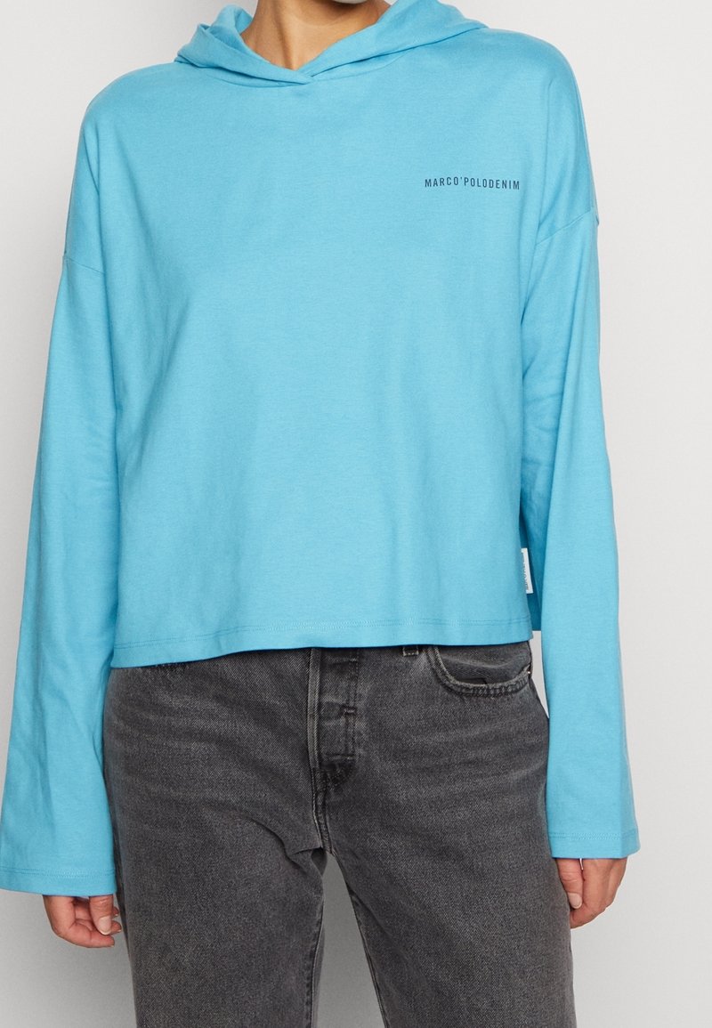 Un hoodie cropped bleu clair en tissu doux, avec des manches longues et un petit logo sur la poitrine, associé à un jean gris.