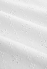 Tissu blanc avec des motifs floraux délicatement brodés et de petites découpes circulaires, présenté avec des plis doux et une texture visible.