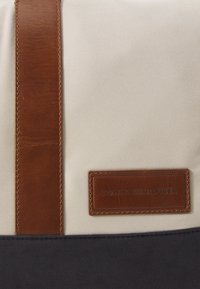 Sac en textile beige avec une bandoulière en cuir marron et une pièce en cuir gravée du logo "TOMMY HILFIGER", et une partie inférieure bleu marine.