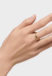 Złoty pierścionek z prostą obrączką i kwiatowym wzorem z grupą zielonych kamieni, noszony na palcu dłoni na jasnym tle.