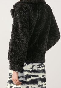 Chaqueta negra de pelaje sintético texturizado con capucha, de corte holgado y mangas amplias, exhibida sobre una falda estampada en tonos de blanco y negro.